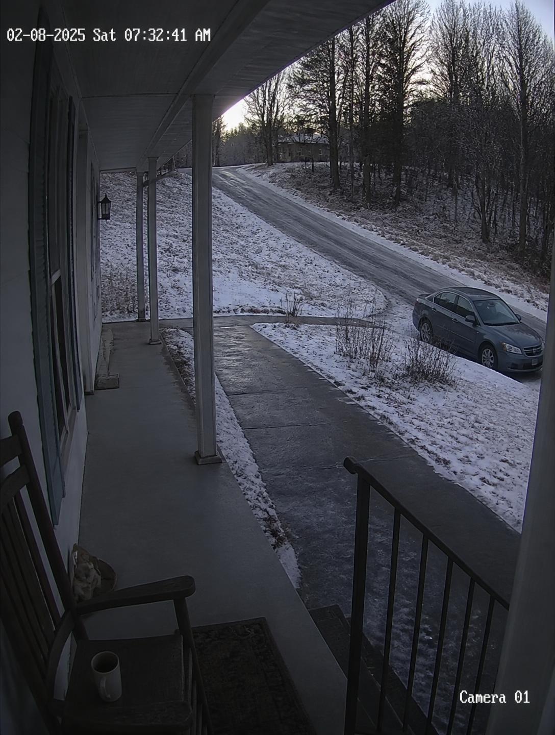 Security camera view of an icy rural front porch, sidewalk, sloping driveway and a parked car in winter